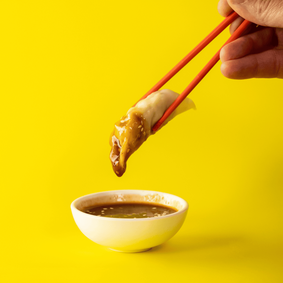 Asian dumpling being dipped into a bowl of sauce with chopsticks on a yellow background